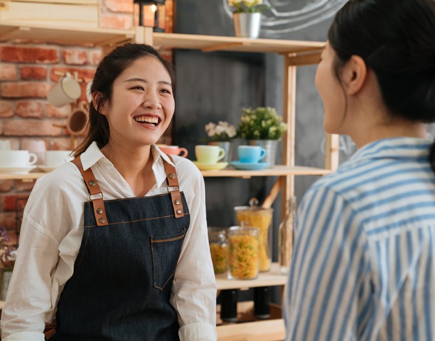 Young barista girl in apron and white shirt working at the counter in coffee shop. Nice smiling coffeehouse female staff taking order from customer in morning. businesswoman is regular client in cafe; Shutterstock ID 1444623287; purchase_order: -; job: -; client: -; other: -