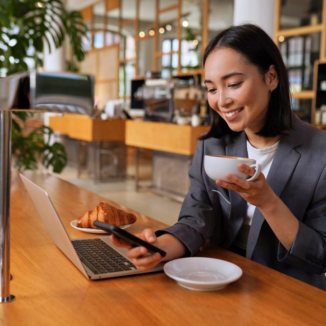 Young Asian business woman wearing suit drinking coffee using smartphone in cafe. Happy smiling female professional working holding mobile phone using smartphone texting messages on cellphone.